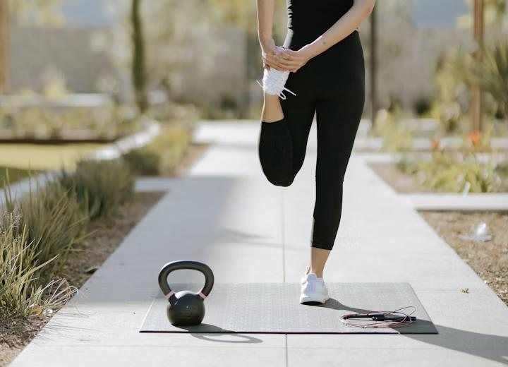 A woman stretching her quad beside a kettlebell and mat outdoors